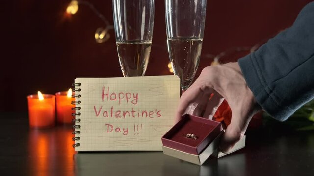 Man Preparing to Make a Marriage Proposal. Putting the Note with Inscription Happy St Valentines Day and Golden Ring on the Table near Glasses with Champagne and Bouquet of Roses