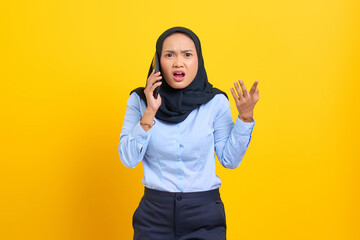 Portrait of surprised young Asian woman talking on mobile phone with open mouth isolated on yellow background