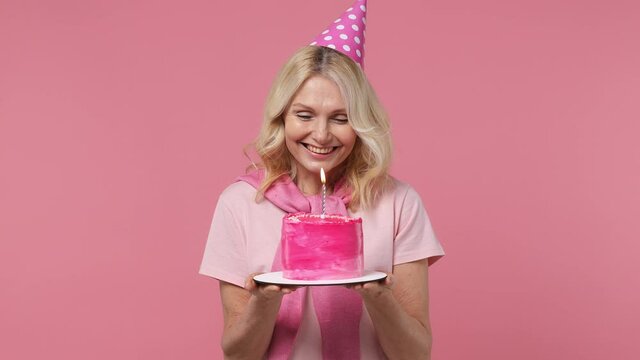Fascinating jubilant excited elderly blonde woman lady 40s years old wears t-shirt birthday hat look camera hold cake with candle smiling isolated on plain pastel light pink background studio portrait