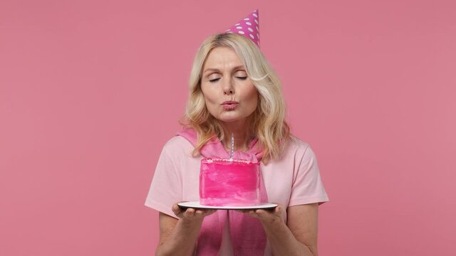 Beautiful cheerful excited fun elderly blonde woman lady 40s years old wears t-shirt birthday hat look camera hold cake blow out candle isolated on plain pastel light pink background studio portrait
