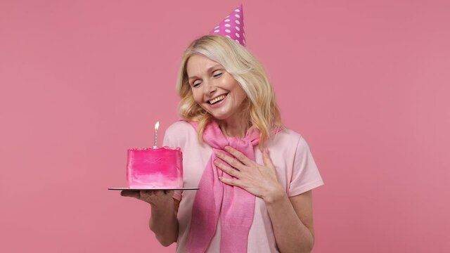 Surprised elderly blonde woman 40s years old wear t-shirt birthday hat hold cake with candle ask for me oh it so sweet put hands on chest isolated on plain pastel light pink background studio portrait