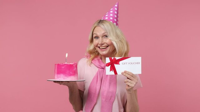 Fun elderly blonde woman lady 40s years old in t-shirt birthday hat hold cake with candle gift certificate coupon voucher card for store isolated on plain pastel light pink background studio portrait