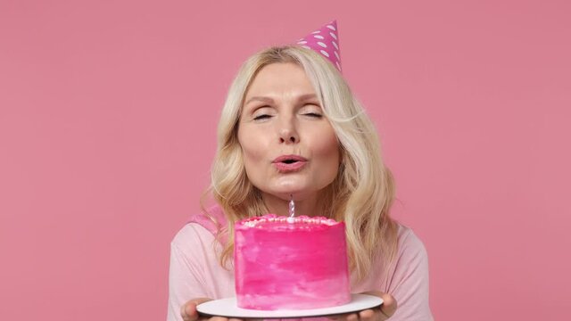 Close up blithesome charming fun elderly blonde woman lady 40s years old wears t-shirt birthday hat look camera hold cake blow out candle isolated on plain pastel light pink background studio portrait