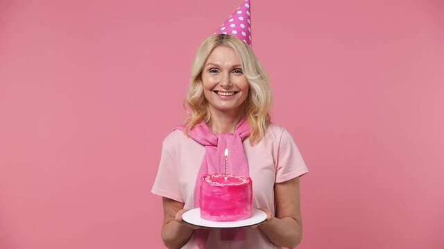 Fancy excited elderly blonde woman lady 40s years old wears t-shirt birthday hat hold cake with candle look at confetti blow out candle isolated on plain pastel light pink background studio portrait