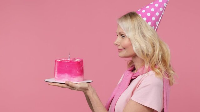 Side view profile charming fun magnificent elderly blonde woman lady 40s years old wears t-shirt birthday hat hold cake blow out candle isolated on plain pastel light pink background studio portrait