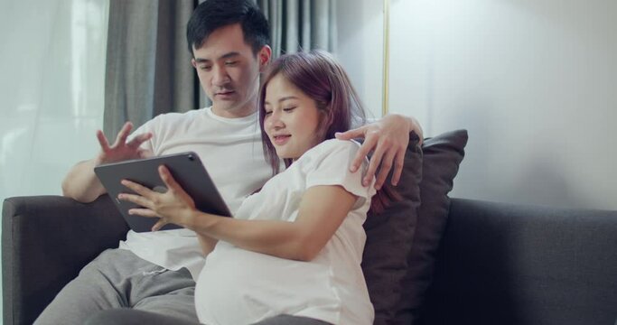 Happy caucasian husband with a pregnant wife looking ultrasound photo of her newborn baby together on a tablet and shopping online in the bedroom at home.