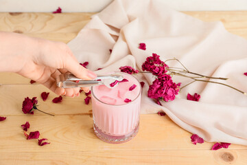 Woman adding ice to tasty rose latte in glass on table
