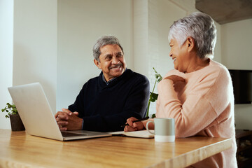 Multi-cultural elderly couple smiling at each other. Sitting at modern kitchen counter with laptop.
