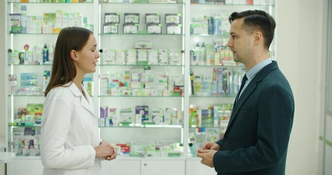 Consultation. Caucasian female young pharmacist druggist advising medicines, pills, drugs, painkillers, vitamins to a male customer client, buyer in drugstore. High quality 4k footage