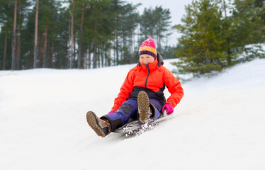 Fototapeta na wymiar childhood, sledging and season concept - happy little girl sliding down on snow saucer sled outdoors in winter over snowy park or forest background