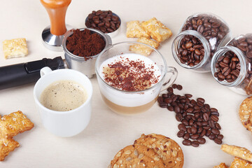 Still life with cup of espresso, cappuccino with chocolate crumbs, cookies, crackers, holder with ground coffee, tamper and coffee beans on table.