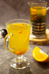 Yellow hot drink in a glass cup on a dark concrete background with a teapot. Sea buckthorn tea with orange.
