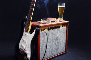 Amplifier for guitar with black guitar, glass of beer and smoking cigarette on black background.