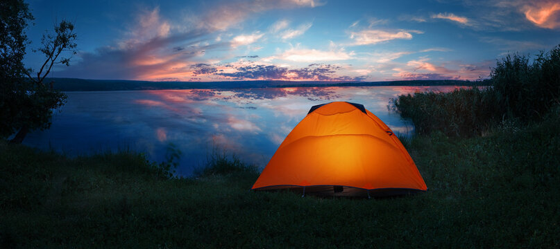 Internally lit orange tent on shore of lake under dramatic sunset sky - a tent pitched tent pitched 