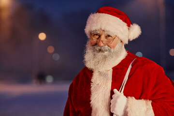 Horizontal medium close-up portrait of cheerful Santa Claus wearing white and red outfit with eyeglasses standing outdoors in evening looking at camera