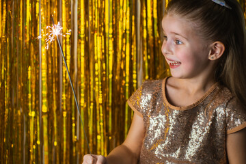 Cute blonde toddler girl wearing fancy hold burning sparkler against golden glitter background and emotionally smile