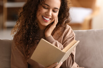 Happy young African-American woman reading book on sofa at home