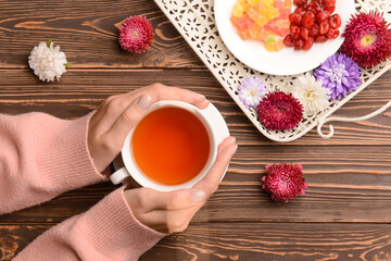 Female hands with cup of tea, sweets and aster flowers on wooden background, closeup