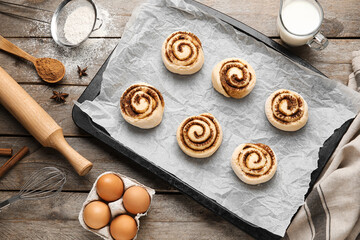 Baking dish with uncooked cinnamon rolls and ingredients on wooden background