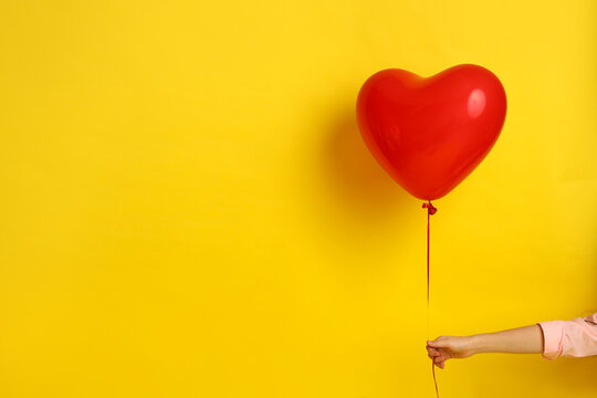 A hand in a pink shirt holds a heart-shaped ball on a yellow background. Picture for Valentine's Day. The concept of gifts and lovers. High-quality photography