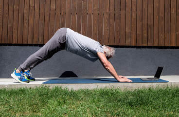 A sporty gray-haired man does yoga online using a computer, performs complex asanas