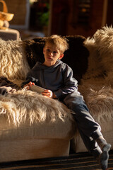 A blonde-haired child is sitting on a cozy sofa in a country house holding a smartphone in his hands