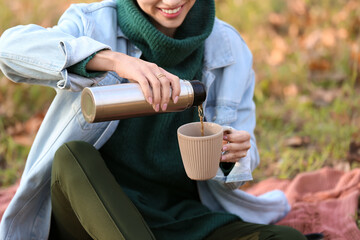 Young woman sitting on plaid and pouring tea from thermos in autumn park