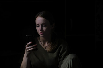 A sad girl is focused on the phone, sits and searches and studies something on the Internet. Portrait of people in a dark key. Depression and addiction