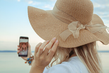 Portrait of girl taking selfie on the smartphone. Young beautiful 30 years woman caucasian appearance in straw hat with big satin ribbon bow. Summer lifestyle concept. Nature background. Pretty face