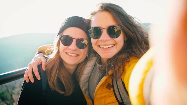 Two young women in sunglasses taking a selfie while standing on an observation deck against the blue sky on a sunny day.