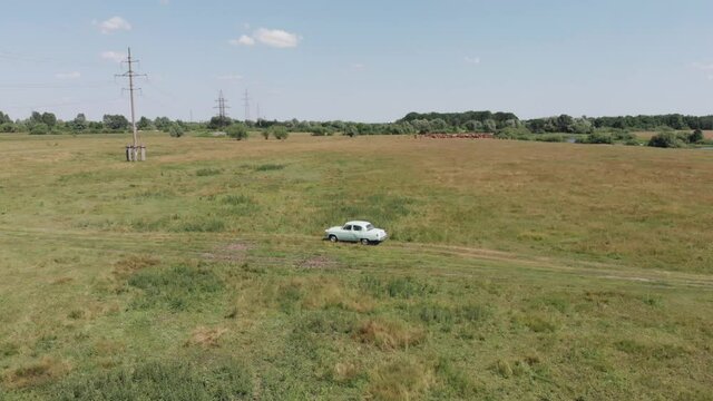 Aerial view of a retro car volga gaz 21 on a well-worn track in a field near a river and a power line on a summer day