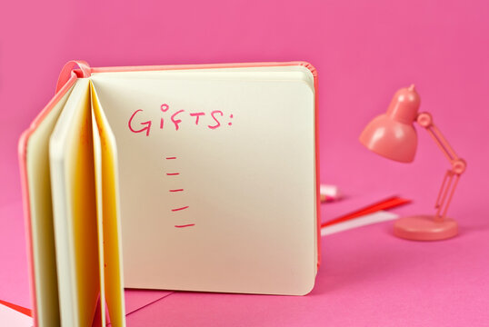 Notebook on a pink background. Toy table lamp near the notebook. Copy space and free space for text in notebook. Mockup for design. The word "gifts" written in pencil. List of gifts for the holidays.