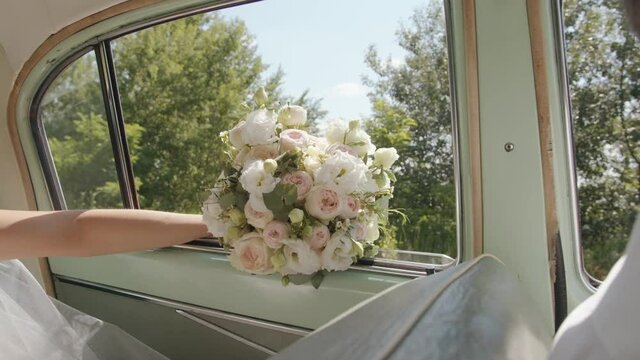 The bride holds a gorgeous wedding bouquet with her hand on the open window of a car driving in nature. Slow motion