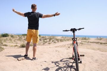 Man standing on beach near bicycle with his arms wide apart back view