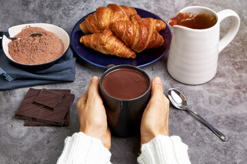 Woman warming her hands with a cup of hot chocolate along with some delicious croissants and various toppings