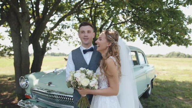 Belarus. Gomel region. August - 21, 2021: Happy groom hugs his laughing bride with a bouquet while standing near a retro car under a tree