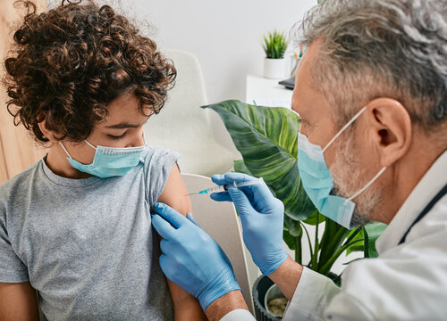 Routine vaccinations for a child. Curly haired boy wearing medical mask while vaccinated by mature pediatrician at medical clinic