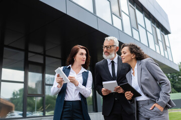 real estate agent showing digital tablet to interracial business partners with notebooks near building