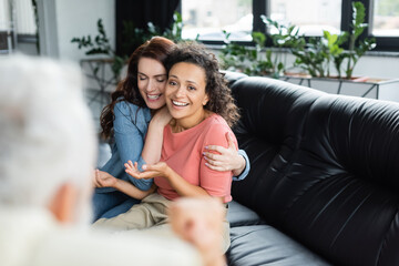 happy lesbian woman embracing african american girlfriend while sitting on sofa near psychologist on blurred foreground