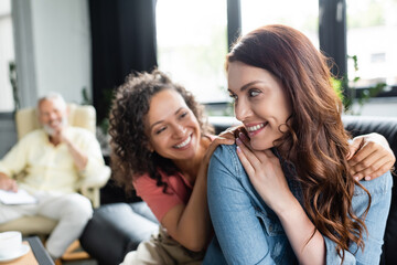 multiethnic lesbian couple smiling near blurred psychologist in consulting room