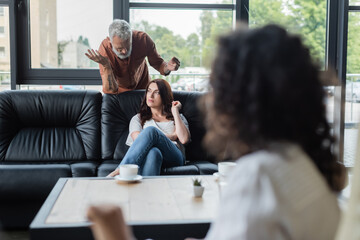 middle aged man gesturing while talking to offended wife during visit to psychologist on blurred...