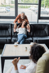 happy man embracing smiling wife sitting on sofa in front of blurred african american psychologist - obrazy, fototapety, plakaty