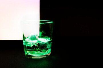 A vessel, a glass half filled with a clear liquid with ice cubes and illuminated with green light on a dark background with a light fragment in the corner