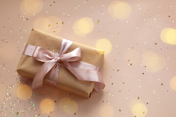 Fototapeta premium Birthday or New Year's eve present in kraft paper with pink ribbon on a soft beige background with glitter and confetti. Xmas composition. Flat lay. Happy holidays celebration and giving love concept