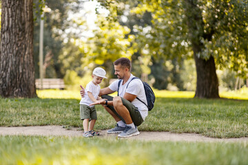 Father and son in nature. Dad squats next to the boy and explains something to him while they are in the woods on a sunny day. Dressed in the same casual clothes, playing in the park, pure happiness