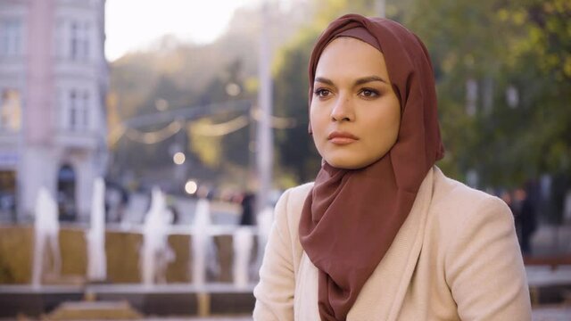 A young beautiful Muslim woman thinks about something in a street in an urban area - a fountain in the blurry background - closeup