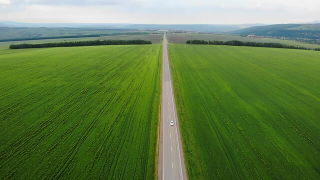 Aerial drone view: white car driving through road among agriculture fields and green countryside, drone following the car.