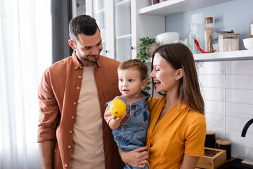 Positive parents looking at toddler son with lemon in kitchen