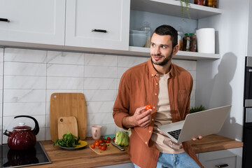 Smiling man holding laptop and cherry tomato in kitchen