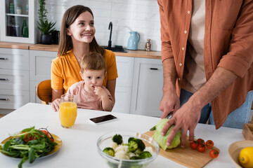 Cheerful woman looking at husband cooking salad near son and smartphone at home